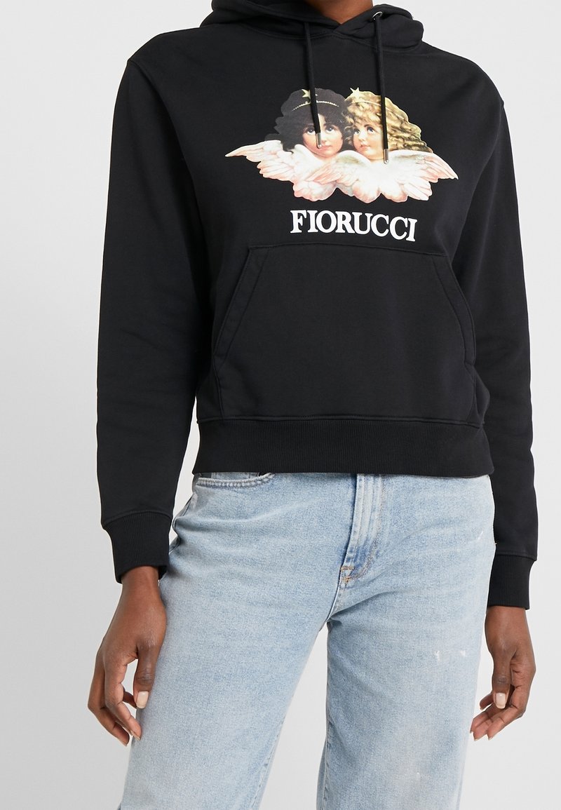 Personne portant un sweat à capuche noir Fiorucci avec un motif de jumeaux anges et un jean bleu clair sur un fond clair uni.