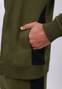Olivenfarbene Zip-Jacke aus weichem Stoff, mit einer seitlichen Tasche mit schwarzem Akzent und gerippten Bündchen am Saum und an den Ärmeln.