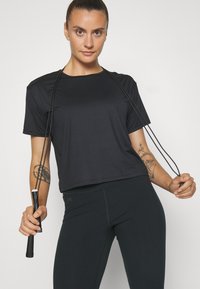 Svart kortermet treningsskjorte med løs passform, kombinert med svarte tights med detaljerte sømmer. En hoppetau holdes i begge hender.