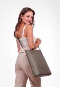 Olivově zelená kožená kabelka obdélníkového tvaru, s vrchním zipem a nastavitelným popruhem, kterou drží osoba oblečená v volné blůze.