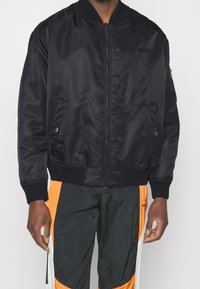 Giubbotto bomber nero con chiusura a zip, polsini e orlo a coste, e logo ricamato "HUGO". Indossato sopra pantaloni sportivi multicolori.
