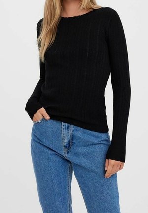 Femme portant un pull noir à manches longues en maille torsadée et un jean bleu taille haute, une main dans la poche, sur un fond uni.