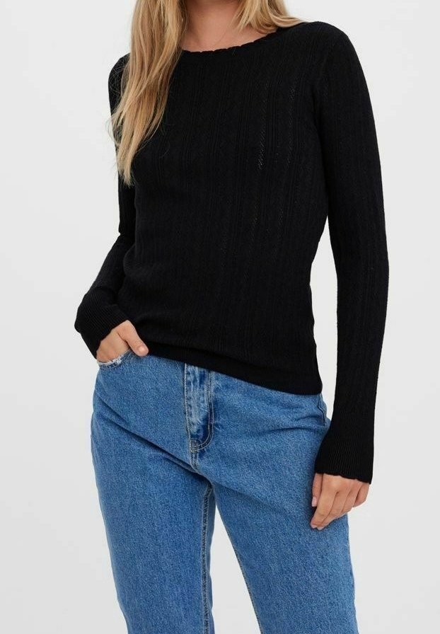 Femme portant un pull noir à manches longues en maille torsadée et un jean bleu taille haute, une main dans la poche, sur un fond uni.