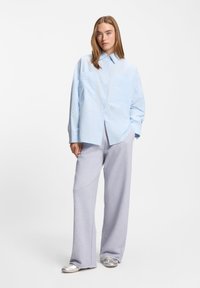 Νέα γυναίκα που φοράει φαρδιά ανοιχτό μπλε πουκάμισο, φαρδιά γκρι παντελόνια και ασημένια αθλητικά παπούτσια, στέκεται με το ένα χέρι στην τσέπη.