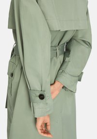 Light green trench coat with a belted waist, buttoned cuffs, and side pockets. Smooth fabric with a minimalist design and structured silhouette.