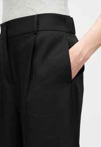 Άτομο που φοράει μαύρο, ραμμένο παντελόνι με το χέρι στην πλαϊνή τσέπη, συνδυασμένο με λευκό ριμπ τοπ, εμφανιζόμενο από τη μέση έως το μέσο του μηρού.