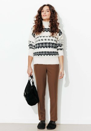 Femme aux cheveux bouclés portant un pull à motif blanc et noir, un pantalon marron, des chaussures noires, tenant un sac à main noir, debout contre un mur blanc.