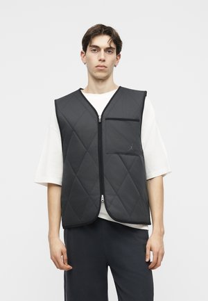 Νέος άνδρας που φοράει μαύρο γιλέκο με επένδυση χωρίς μανίκια, με φερμουάρ μπροστά και τσέπη στο στήθος πάνω από ένα λευκό κοντομάνικο πουκάμισο.