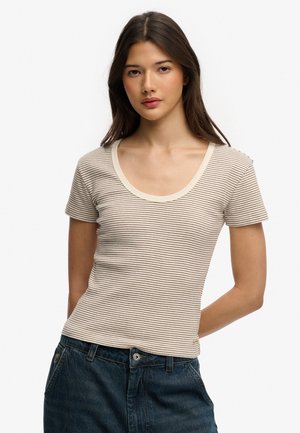 Ung kvinde med langt mørkt hår iført en beige og hvidstribet kortærmet skjorte og mørke cowboybukser, stående mod en hvid baggrund.