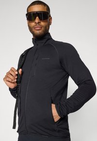 Svart prestasjonsjakke med høy krage, strukturert stoff, glidelås-lomme på brystet, to sidelommer og snug passform, kombinert med oversized solbriller.