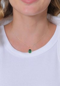 Guld kædedenkæde med en rund, grøn ædelsten vedhæng, båret over en hvid ribbet top. Designet er simpelt og elegant.