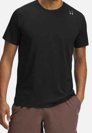 Homme portant un t-shirt de sport noir à manches courtes avec un petit logo sur l'épaule et un short marron, main dans la poche.
