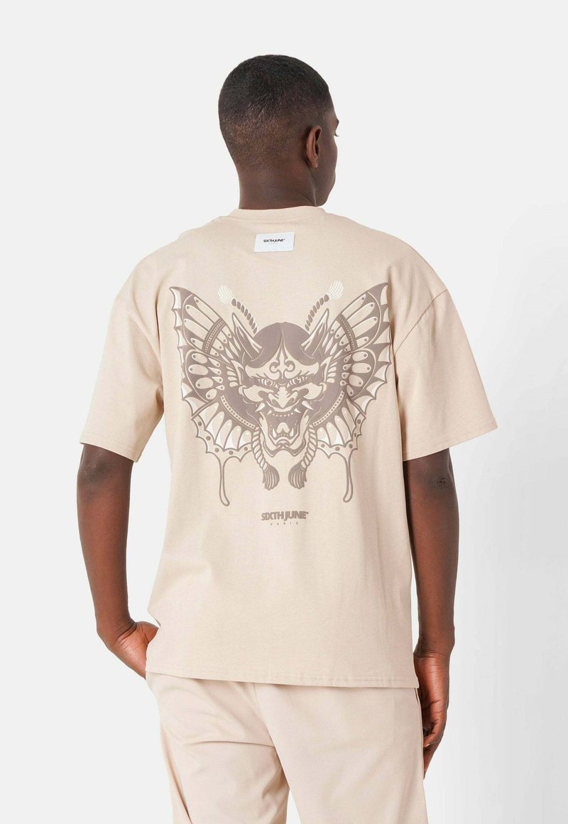 Camiseta de algodón beige con un gran gráfico gris de una máscara y alas de mariposa en la espalda, con mangas cortas y un corte holgado.