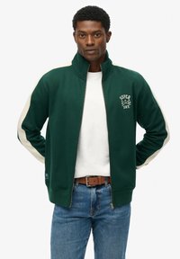 Grøn zip-up sweatshirt med hvide side-striber, lavet af bomulds materiale, høj krave og et Superdry logo på brystet, kombineret med denim jeans.