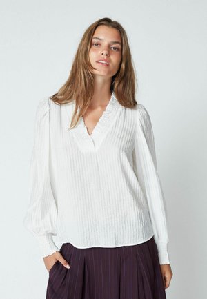 Blusa bianca con texture, scollatura a V e rifiniture arricciate. Maniche lunghe a palloncino con polsini arricciati. Abbinata a pantaloni scuri con pinces.
