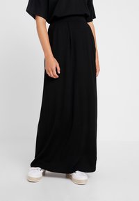 Black maxi skirt made of soft, flowing fabric, featuring pleats at the waist. Paired with white espadrille-style shoes for contrast.