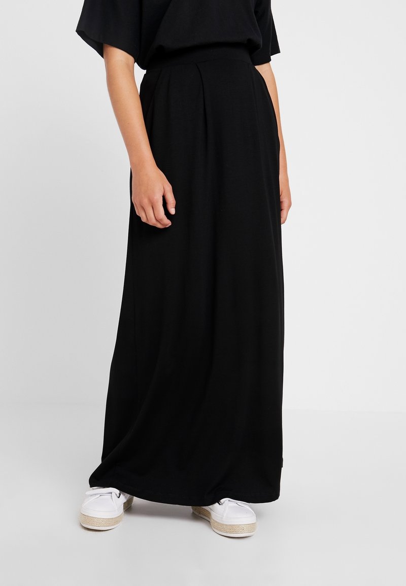 Black maxi skirt made of soft, flowing fabric, featuring pleats at the waist. Paired with white espadrille-style shoes for contrast.