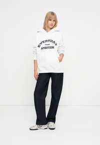 Felpa bianca con cappuccio e testo blu navy "EVERYDAY AMBITION", tasca frontale, abbinata a jeans a gamba larga blu scuro e sneaker con motivo.