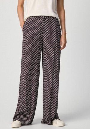 Persona vistiendo pantalones negros sueltos con un patrón floral blanco y rosa y zapatillas blancas, de pie con las manos en los bolsillos.