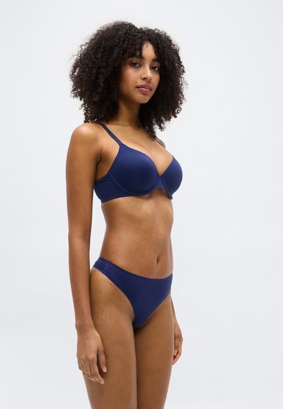 Γυναίκα με σγουρά μαλλιά που φοράει σετ σουτιέν και εσώρουχα σε navy blue χρώμα, στεκόμενη πλευρικά μπροστά σε απλό ανοιχτό φόντο.