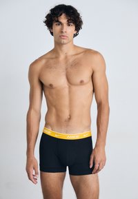 Boxer briefs negros para hombres con un logo de Nike amarillo en la cinturilla. La tela parece suave y tiene un diseño ajustado.
