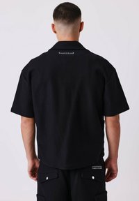 Maglietta nera a maniche corte con colletto, caratterizzata da una finitura testurizzata e un'etichetta di marca sul retro, indossata con pantaloni abbinati.