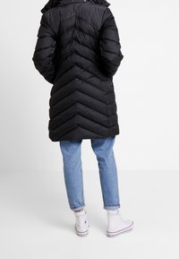 Schwarzer Steppmantel mit Chevron-Steppung, der bis zur Mitte des Oberschenkels reicht, kombiniert mit hellblauen aufgekrempelten Jeans und weißen High-Top-Sneakers.