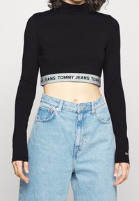 Black long-sleeve crop top with mock neck and gray elastic waistband featuring "TOMMY JEANS" text. Blue high-waisted denim jeans.