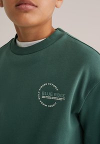 Hanorac verde din bumbac, cu un imprimeu rotund al logo-ului și text alb, guler ribat și design lejer.