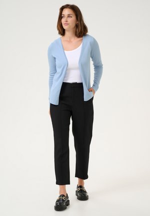 Gilet bleu clair, à manches longues, ouvert sur le devant. Débardeur blanc en dessous. Pantalon noir ajusté à coupe droite. Mocassins noirs avec des accents dorés.
