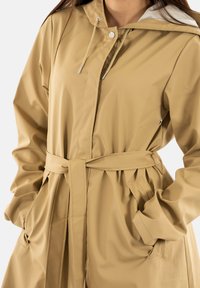 Beige waterdichte regenjas met een getailleerde taille, capuchon en drukknoop/sluiting. Voorzien van zakken en een gladde, licht glanzende textuur.