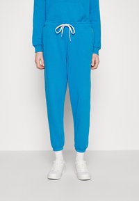 Person klädd i ljusblåa sweatpants med vit dragsko, vita strumpor och vita sneakers stående mot en enfärgad bakgrund.