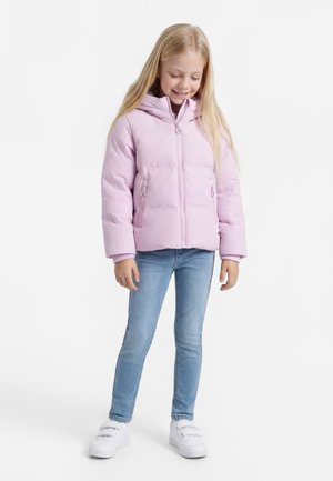 Niña joven con pelo largo y rubio que lleva una chaqueta acolchada morado claro, vaqueros azul claro y zapatillas blancas, de pie y mirando hacia abajo.