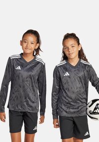 Graue Langarm-Fußballtrikots mit abstraktem Muster, weißen Akzenten und V-Ausschnitt; kombiniert mit schwarzen Shorts. Eine Person hält einen Fußball.