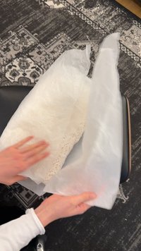 Handen die een wit stuk stof met kanten rand vouwen op een zwarte stoel bovenop een patroonrijke zwart-witte kleed.