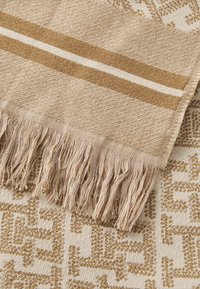 Tappeto in cotone beige intrecciato con accenti dorati, caratterizzato da bordi frangiati e motivi geometrici. Texture morbida e forma rettangolare.