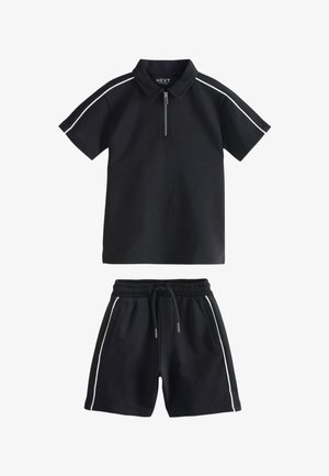 Crna polo majica s kratkim rukavima i prednjim patentnim zatvaračem te crne kratke hlače s elastičnim strukom, oba s bijelim prugama sa strane.