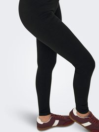 Gambe indossando leggings neri aderenti e scarpe da ginnastica color amaranto con dettagli bianchi, in piedi su uno sfondo grigio.