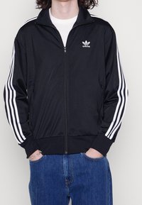 Svart zip-up träningsjacka med vita tre-randiga ärmar och Adidas-logotypen på bröstet. Tillverkad av ett smidigt, lätt material.