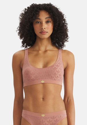 Frau mit lockigem Haar, die einen passenden rosa Sport-BH und Slips mit dezenten Blattmustern und kleinen gelben adidas-Logos am Bund trägt.