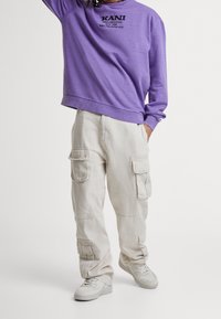 Felpa viola con testo nero, abbinata a pantaloni cargo beige chiaro caratterizzati da numerose tasche e un fit rilassato. Le sneakers bianche completano l'outfit.