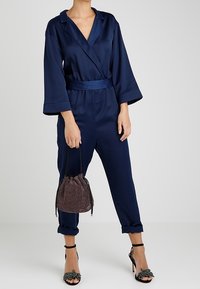 Navy blue, satin jumpsuit with wide sleeves, belted waist, cuffed legs. Paired with a sparkly drawstring bag and strappy high-heeled sandals.