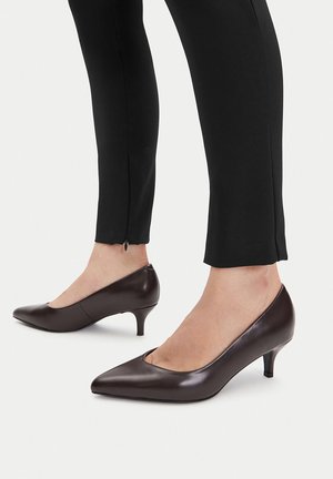 Brązowe skórzane czółenka na wysokim obcasie z ostrym noskiem, posiadające obcas o średniej wysokości i gładką fakturę, zestawione z czarnymi dopasowanymi spodniami.