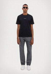 Mann iført svart Michael Kors T-skjorte, grå jeans, hvite sneakers og svarte solbriller som står foran en enkel hvit bakgrunn.