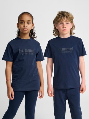 Zwei Kinder stehen nebeneinander und tragen passende dunkelblaue hummel Sport-T-Shirts und Hosen vor einem einfarbigen Hintergrund.