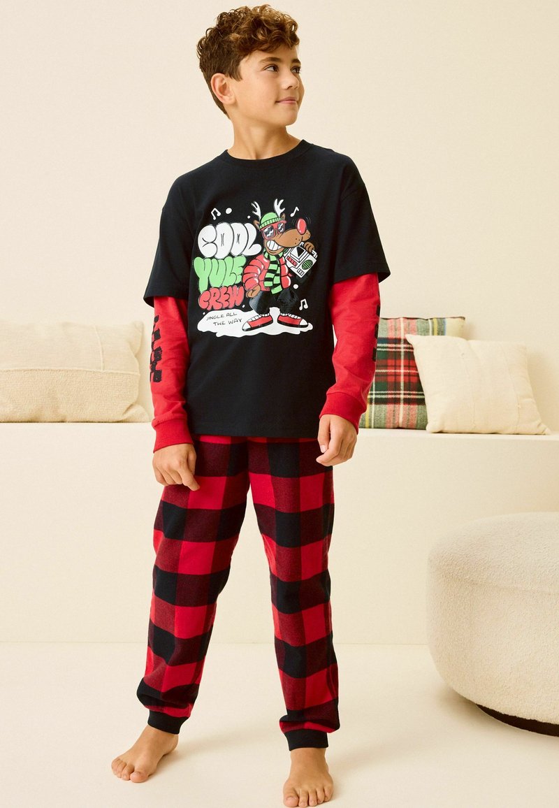 Czarny t-shirt z krótkim rękawem z grafiką przedstawiającą renifera, czerwone długie rękawy pod spodem, czerwono-czarną w kratkę piżamę.