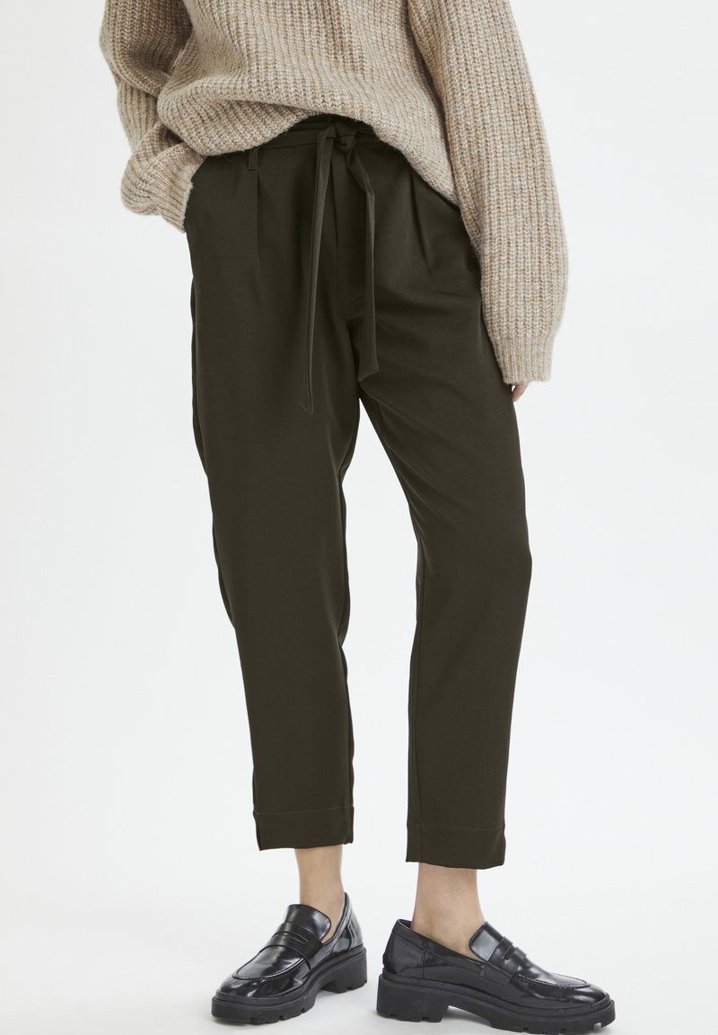 Un pantalon taille haute vert foncé avec une ceinture à nouer, des jambes fuselées et des accents cousus, porté avec des mocassins en cuir noirs.