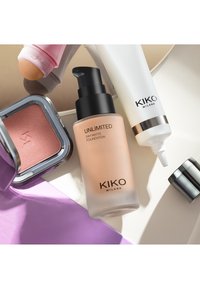 KIKO Milano Unlimited matte foundation i en frostet glassflaske med en svart pumpe, omgitt av sminkeprodukter i ulike former og farger.