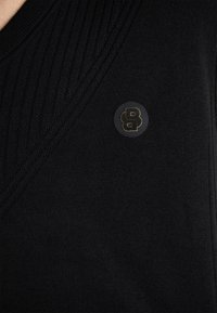 Czarny sweter z prążkowanym dekoltem w kształcie litery V. Posiada okrągłą naszywkę z logo w stylizowanej wersji litery 'B' w kolorze złotym na przodzie. Gładka tekstura.