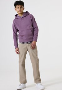 Un chico adolescente de pie, vestido con una sudadera morada, pantalones beige, zapatillas blancas y una camisa de capas debajo, contra un fondo blanco liso.